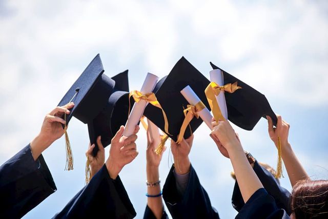 Studenter i akademiska dräkter höjer sina diplom och hattar mot en blå himmel.